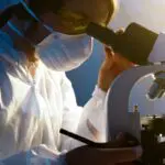 A scientist wearing protective clothing, a mask, and safety glasses looks through a microscope in a laboratory setting, with a pen in hand and focused on her research.
