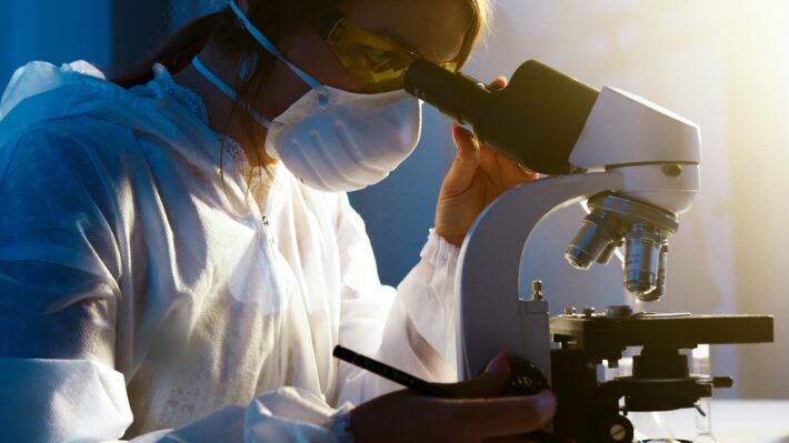 A scientist wearing protective clothing, a mask, and safety glasses looks through a microscope in a laboratory setting, with a pen in hand and focused on her research.