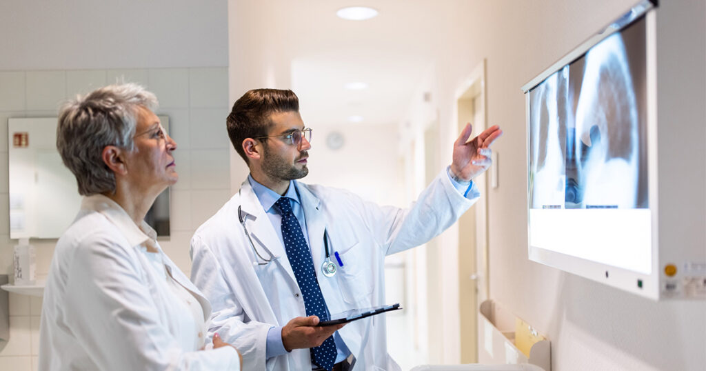A male doctor in a white coat points at chest X-rays on a screen while explaining them to a female colleague in a medical setting. The doctor holds a tablet in his other hand.