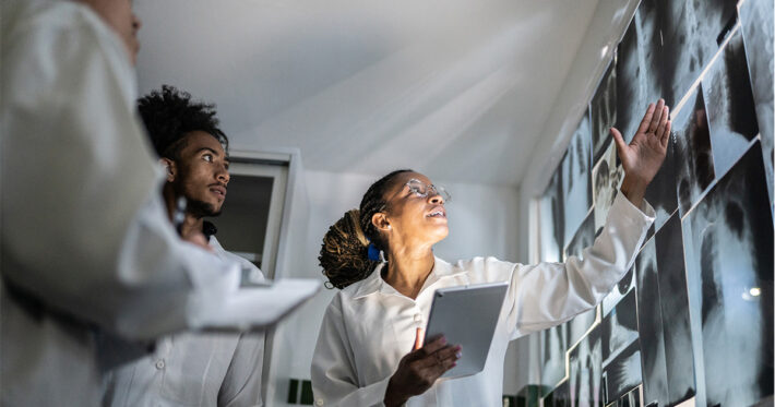 A group of medical professionals in lab coats examines and discusses X-ray images on a wall. One person points at the images while holding a tablet, and others listen and take notes.