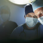Two surgeons wearing surgical masks and caps focus intently on a procedure under bright operating room lights, with another medical professional visible in the background.