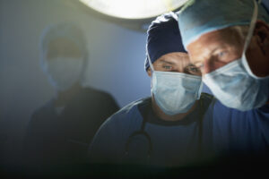 Two surgeons wearing surgical masks and caps focus intently on a procedure under bright operating room lights, with another medical professional visible in the background.