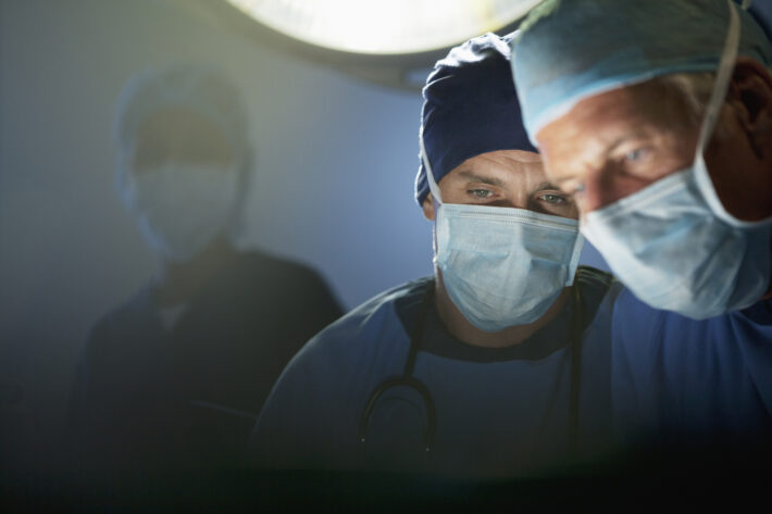 Two surgeons wearing surgical masks and caps focus intently on a procedure under bright operating room lights, with another medical professional visible in the background.