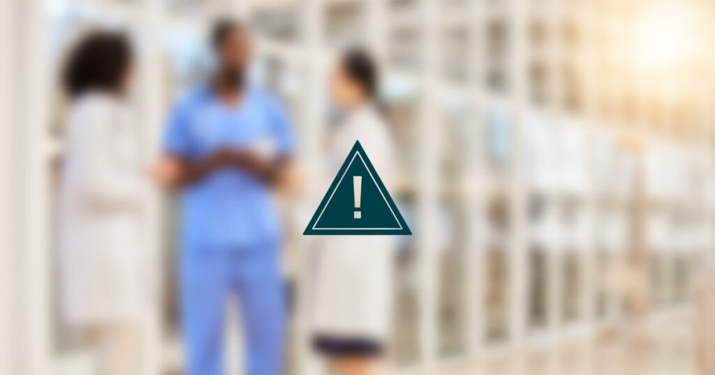 Three people in medical attire are standing and talking in a brightly lit hallway. The image is blurred, and there is an exclamation mark inside a triangle warning icon overlayed in the center.
