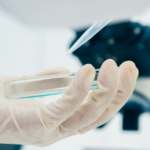 A gloved hand holds a clear petri dish near a microscope in a bright laboratory setting, suggesting scientific research or experiment.