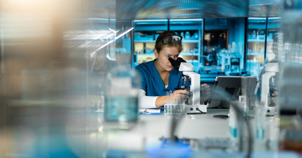 A scientist in blue scrubs looks into a microscope in a modern laboratory, surrounded by glassware, test tubes, and laboratory equipment with shelves of supplies in the background.