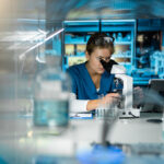 A scientist in blue scrubs looks into a microscope in a modern laboratory, surrounded by glassware, test tubes, and laboratory equipment with shelves of supplies in the background.