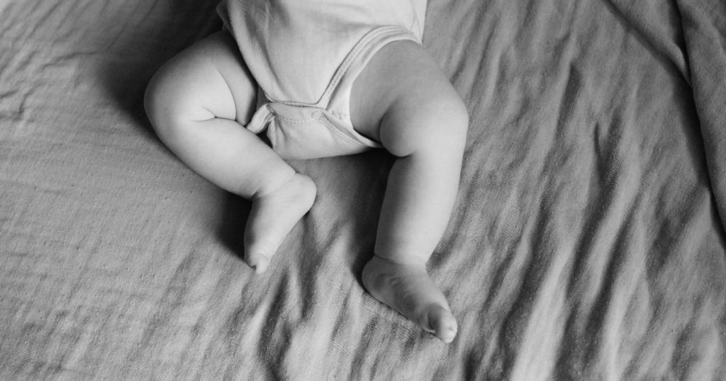 Black and white photo showing a baby lying on a soft, wrinkled blanket. Only the baby’s legs and lower torso are visible, wearing a short-sleeved onesie. The baby’s legs are slightly bent.