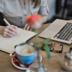 A person writes in a notebook next to an open laptop, a smartphone, a teacup with a saucer, and sugar bowls on a wooden table. A pink flower in a vase is blurred in the foreground.