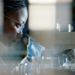 A scientist wearing safety glasses and gloves closely examines samples through a microscope in a laboratory setting, concentrating on her research.