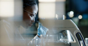 A scientist wearing safety glasses and gloves closely examines samples through a microscope in a laboratory setting, concentrating on her research.