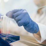 A person in a lab coat and blue gloves is placing a test tube into a blue rack in a laboratory setting, with lab equipment visible in the background.