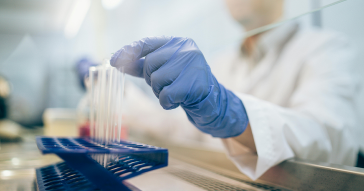A person in a lab coat and blue gloves is placing a test tube into a blue rack in a laboratory setting, with lab equipment visible in the background.
