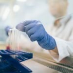 A person wearing blue gloves and a white lab coat places test tubes into a blue rack in a laboratory setting, with scientific equipment visible in the background.