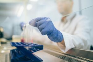A person wearing blue gloves and a white lab coat places test tubes into a blue rack in a laboratory setting, with scientific equipment visible in the background.