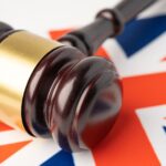 A wooden judge’s gavel with a gold band rests on top of a British flag, symbolizing law or justice in the United Kingdom.