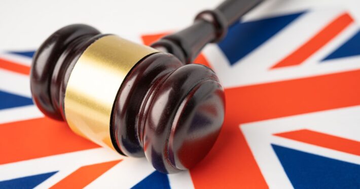 A wooden judge’s gavel with a gold band rests on top of a British flag, symbolizing law or justice in the United Kingdom.