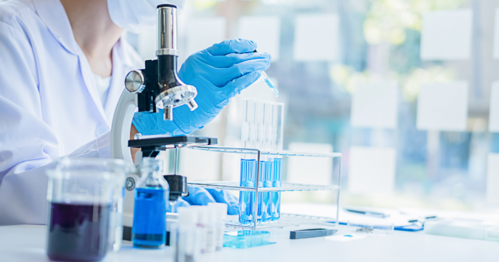 A scientist in a lab coat and gloves uses a pipette to add liquid to a test tube. Several test tubes filled with blue liquid are in a rack beside a microscope on a bright laboratory desk.