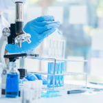 A scientist in a lab coat and gloves uses a pipette to add liquid to a test tube. Several test tubes filled with blue liquid are in a rack beside a microscope on a bright laboratory desk.