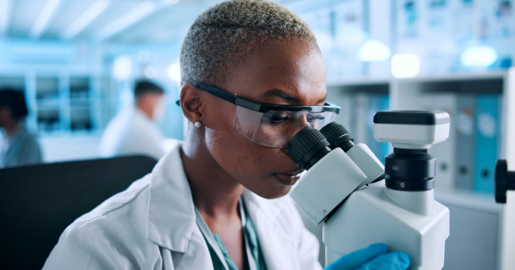 A scientist wearing safety goggles and gloves examines samples through a microscope in a bright laboratory, with other researchers working in the background.
