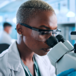 A scientist wearing safety goggles and gloves examines samples through a microscope in a bright laboratory, with other researchers working in the background.