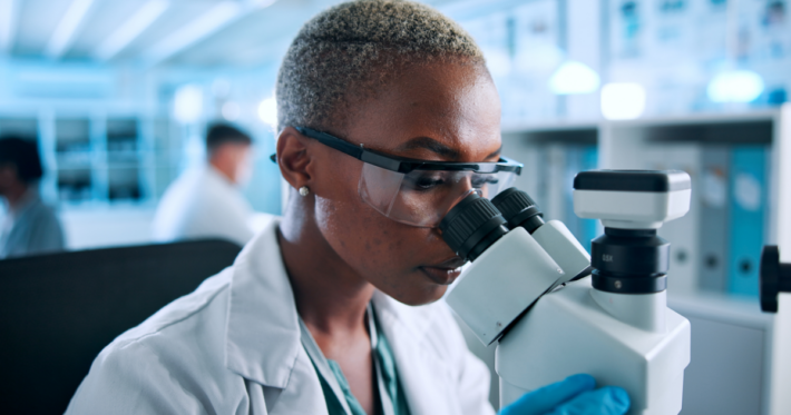 A scientist wearing safety goggles and gloves examines samples through a microscope in a bright laboratory, with other researchers working in the background.