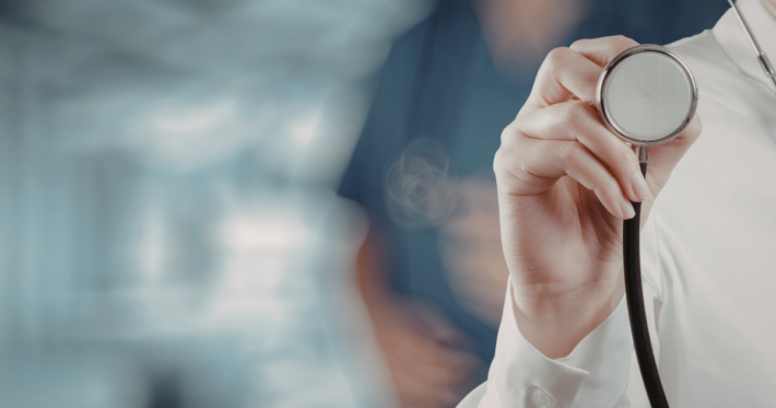 A doctor in a white coat holds a stethoscope toward the camera, with a blurred medical professional in blue scrubs in the background. The setting appears to be a hospital or clinic.
