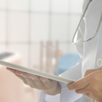 A doctor in a white coat with a stethoscope around their neck holds a tablet device, standing in a medical setting with a patient lying on a bed in the background.