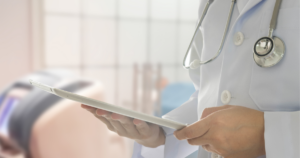 A doctor in a white coat with a stethoscope around their neck holds a tablet device, standing in a medical setting with a patient lying on a bed in the background.
