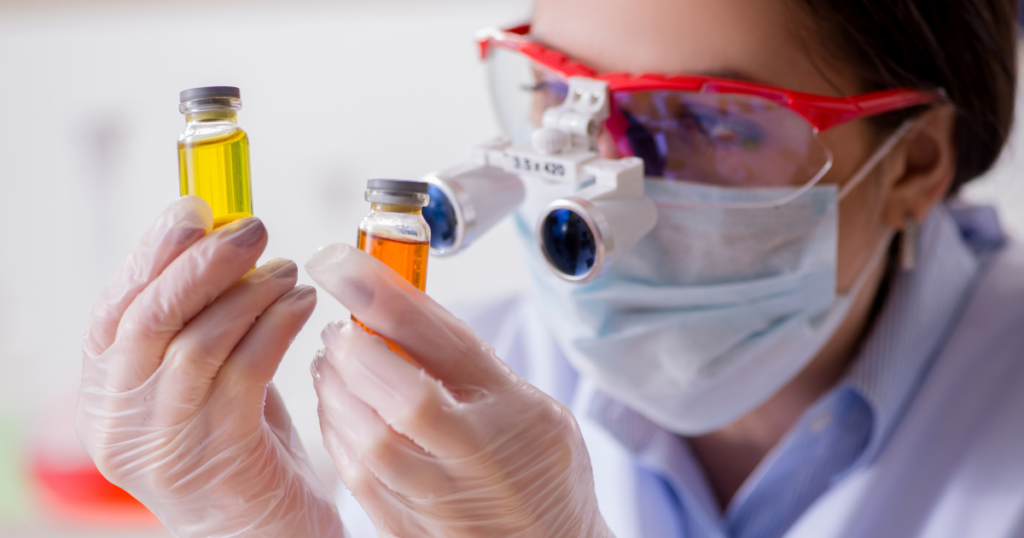 A person wearing protective goggles, face mask, and gloves examines two small glass vials containing yellow and orange liquids in a laboratory setting.