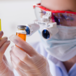 A person wearing protective goggles, face mask, and gloves examines two small glass vials containing yellow and orange liquids in a laboratory setting.