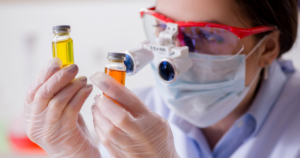 A person wearing protective goggles, face mask, and gloves examines two small glass vials containing yellow and orange liquids in a laboratory setting.