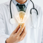 A doctor wearing a white coat and stethoscope holds a glowing light bulb in one hand, symbolizing innovation or new ideas in healthcare.