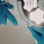A close-up view of two hands in blue surgical gloves passing a surgical instrument under bright operating room lights.