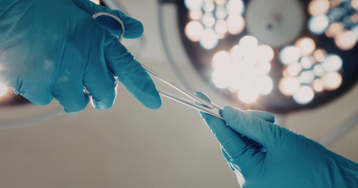 A close-up view of two hands in blue surgical gloves passing a surgical instrument under bright operating room lights.