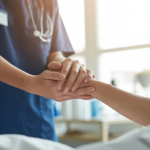 A healthcare professional in blue scrubs holds the hand of a patient in bed, offering comfort and support in a bright, sunlit room.