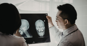 Two doctors examine X-ray images of a human skull, one in profile and one facing forward, while discussing the results in a well-lit medical setting.