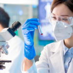A scientist wearing a lab coat, gloves, safety goggles, and a mask examines a test tube beside a microscope in a laboratory. Another scientist works in the background. Scientific equipment and screens are visible.