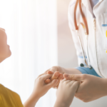 A doctor wearing a stethoscope and a yellow ribbon holds hands with a smiling child dressed in yellow, symbolizing support and care in a bright, sunlit room.
